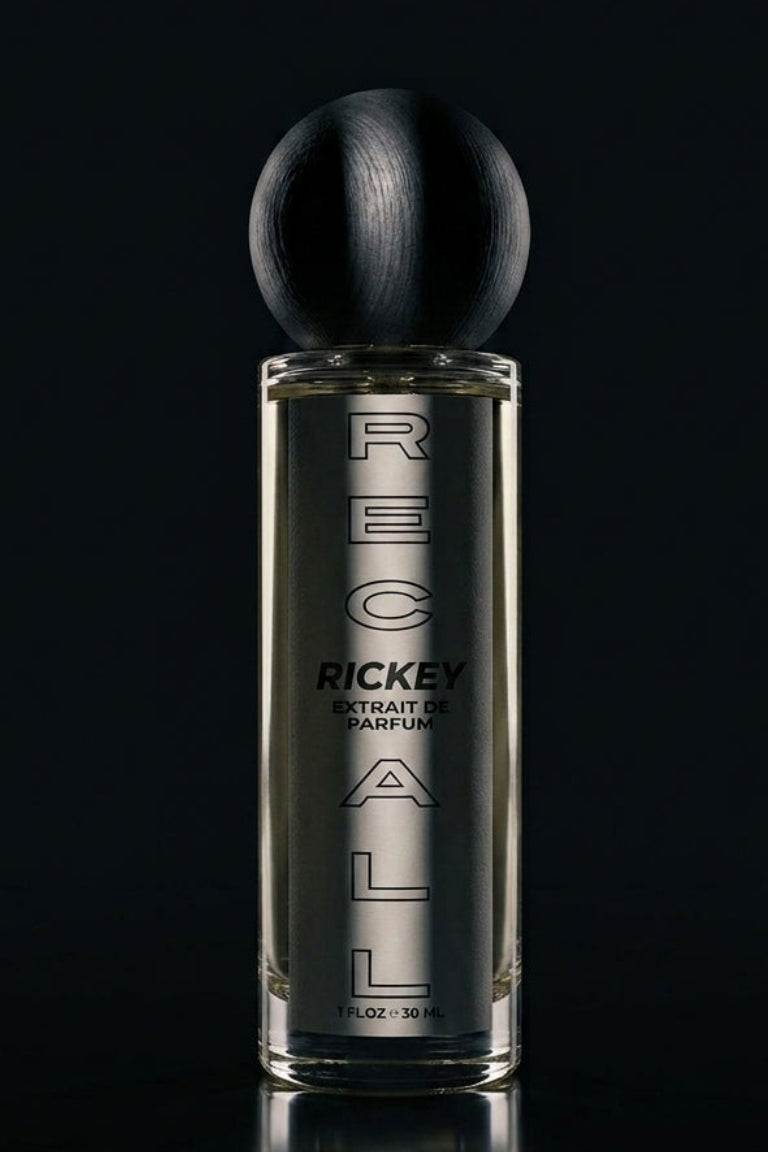 A bottle of Ricky arfum, featuring a clear glass bottle with silver lettering and a black spherical cap, set against a dark reflective background.